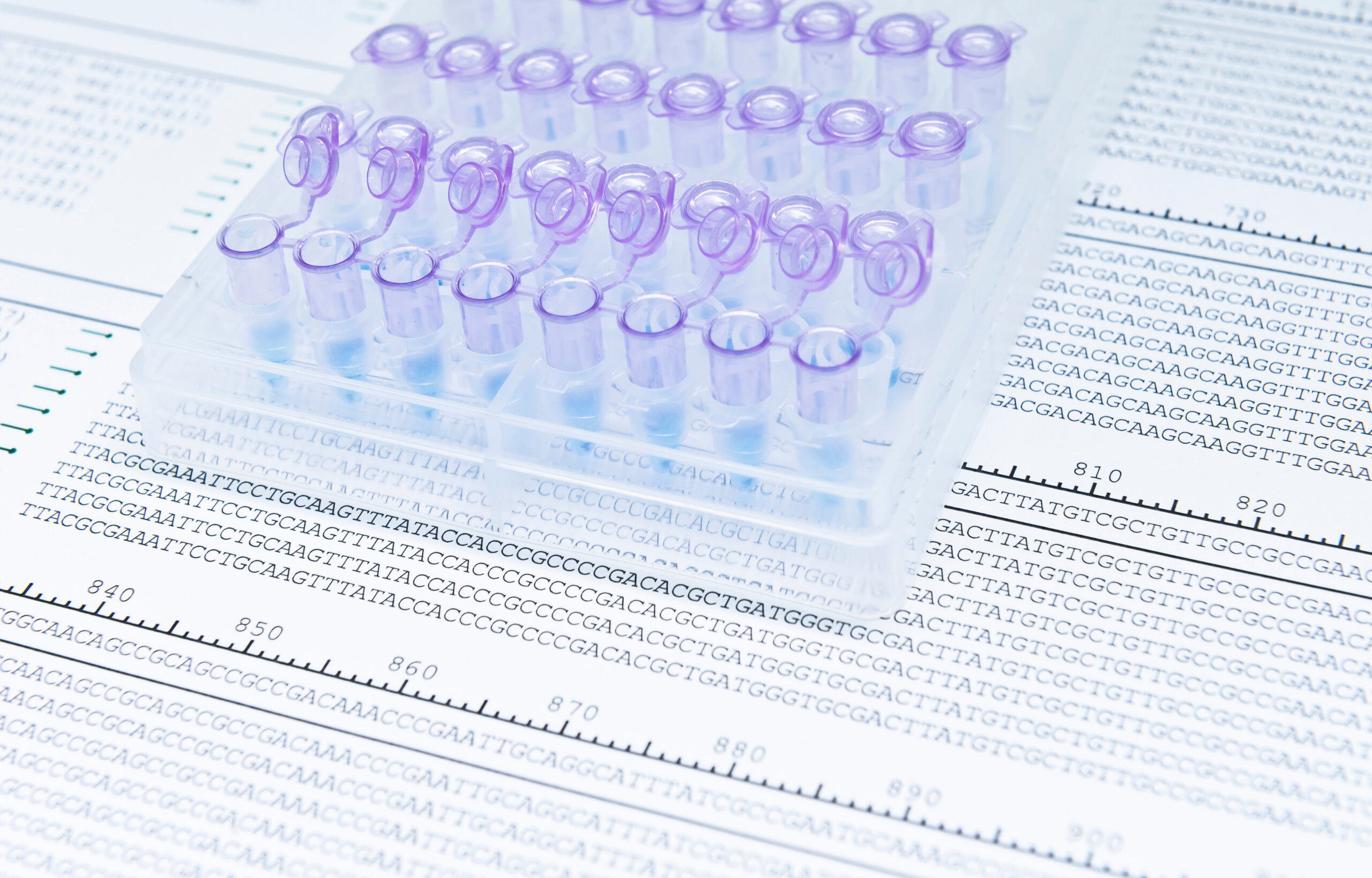Plastic microcentrifuge tubes in a rack placed over printed DNA sequence reads, representing genome sequencing and bioinformatics analysis.